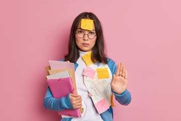 Serious Asian female student does stop gesture keeps palm forward camera asks not to bother her as she prepares for final exams poses with papers sticky notes isolated over pink studio background.