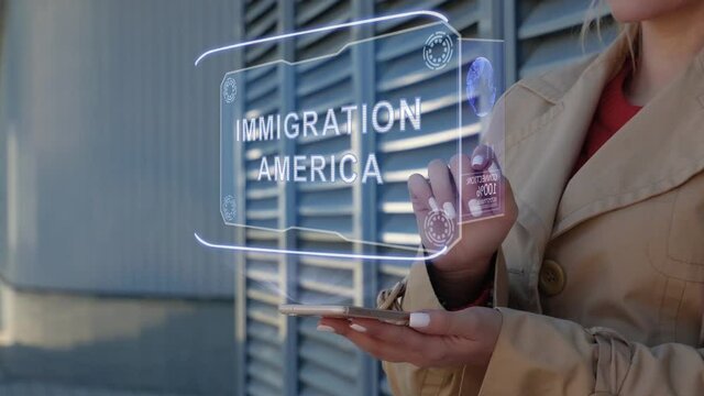 Unrecognizable businesswoman interacts HUD hologram with text Immigration America. Woman in the coat uses the technology of the future mobile screen on the background of the city