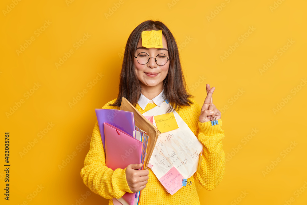 Wall mural pleased asain female student believes in good luck at exam stands with eyes closed and fingers cross