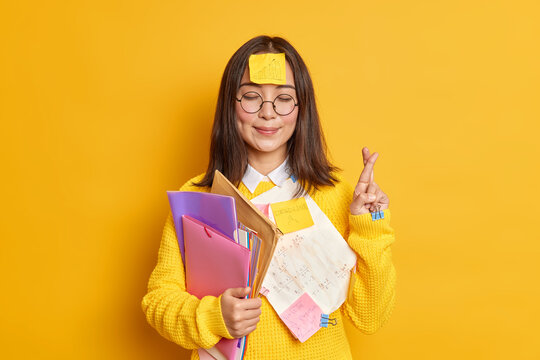 Pleased Asain female student believes in good luck at exam stands with eyes closed and fingers crossed believes dreams come true stuck with papers holds folders isolated over yellow background