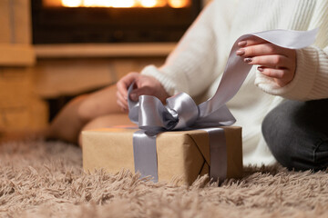 Fototapeta na wymiar A young woman sits on the floor by the fireplace and unties a bow on a box with a gift. The concept of a holiday and surprises.