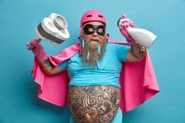 Positive bearded superhero holds cleaning detergent and plunger washes everything on his way busy with domestic chores wears helmet mask rubber gloves and cape isolated over blue background.