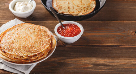 Traditional Russian Crepes Blini stacked in plate and cast-iron frying pan with red caviar, fresh sour cream on dark wooden table. Russian festival meal Maslenitsa or Shrovetide.