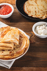 Traditional Russian Crepes Blini stacked in plate and cast-iron frying pan with red caviar, fresh sour cream on dark wooden table. Russian festival meal Maslenitsa or Shrovetide.