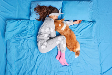 View from above of serene young woman has deep healthy sleep with dog poses in comfortable bed on blue fresh bedclothes wears soft pajama enjoys rest after working day. People coziness sleeping