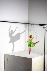 Ballerina made of cabbage, spicy pepper, cucumbers. Flying food composition making beautiful sportswoman drawing shadow on the white wall. Concept of sport, nutrition, healthy lifestyle choice.