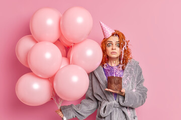 Holidays concept. Shocked embarrassed redhead woman with leaked makeup after crying celebrates birthday alone holds chocolate cake and inflated airballoons wears domestic clothes poses indoor