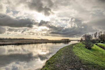 Fototapeta na wymiar Low sun in a dramatic sky over a muddy hiking trail along a canal on a windless day in winter in Vlietlanden nature reserve near Maasland, The Netherlands
