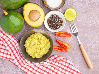 Organic avocado with seed, avocado halves, whole fruit, and fresh guacamole in a bowl with salt and pepper on a small dish on gray stone background. Top view. Concept of healthy fruit