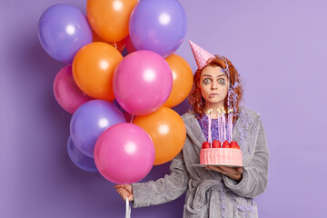 Shocked embarrassed woman with ginger hair cream on head and clothes carries inflated colorful balloons holds strawberry cake wears party hat celebrates special occasion. Puzzled birthday girl - obrazy, fototapety, plakaty