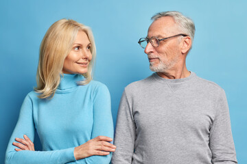 Portrait of self confident blonde woman stands crossed arms and looks happily at her mature husband have pleasant talk together look at each other isolated over blue background. Elderly couple