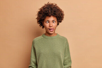 Fototapeta na wymiar Crazy mad dark skinned woman crosses eyes and sticks out tongue makes grimace foolishes around after all day studying wears casual jumper plays fool isolated over brown background. Funny face
