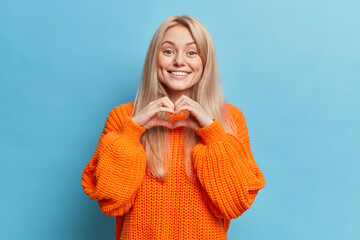 Happy blonde woman shapes heart gesture expresses love smiles pleasantly wears casual knitted...