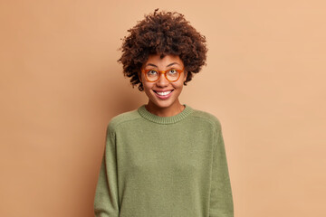 Fototapeta na wymiar Happy carefree African American woman smiles broadly concentrated above with cheerful expression wears casual jumper transparent glasses isolated over beige background. Positive emotions concept