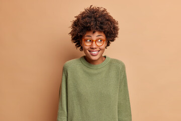 Portrait of pretty positive ethnic woman looks aside with toothy smile sees something nice wears casual long sleeved jumper and spectacles poses in studio against brown background. Good emotions