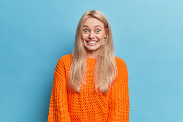 Portrait of surprised blonde young woman smiles broadly reacts happily on awesome news has perfect...