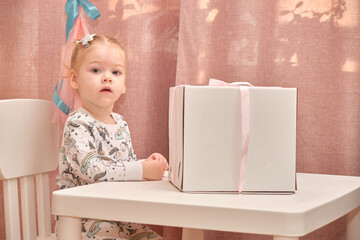 A little girl in pajamas on her birthday sits on a white chair at a children's table, on which there is a birth.