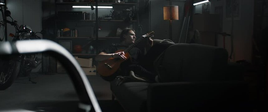 WIDE Caucasian teenager boy lying on a sofa in his home garage hideout, playing guitar. Shot with 2x anamorphic lens