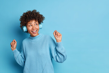 Positive carefree African American woman dances carefree keeps arms raise smiles broadly has fun enjoys favorite music wears wireless headphones dressed in sweater isolated over blue wall copy space