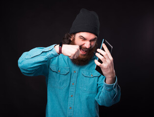 A bearded man is screaming and puching the phone that he is holding near a black wall . © Vulp