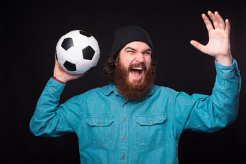 Naklejka premium A young excited bearded man is screaming and holding a soccer ball up near a black wall .