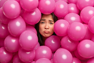 Fototapeta na wymiar Upset displeased miserable Asian woman surrounded with inflated pink balloons has bad mood on holidays because of quarantine purses lower lip. Boring birthday party. Negative emotions concept