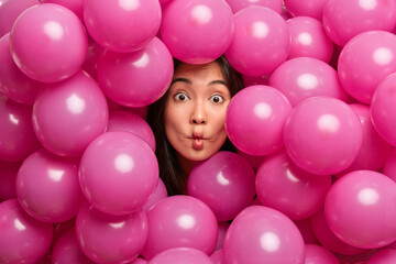 Fototapeta na wymiar Beautiful brunette young Asian woman makes fish lips has funny expression sticks out head through inflated rosy balloons foolishes around at birthday party. Decoration. Special occasion concept