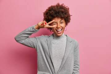 Positive young African American woman has fun wears formal clothes makes peace gesture smiles joyfully poses against pink background. Outgoing optimistic dark skinned female sends good vibes