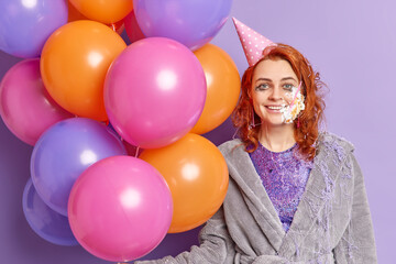 Obraz premium Happy beautiful redhead young woman has fun on birthday party wears cone hat smeared with cream holds colorful balloons celebrates special occasion at home dressed in soft robe. After holiday