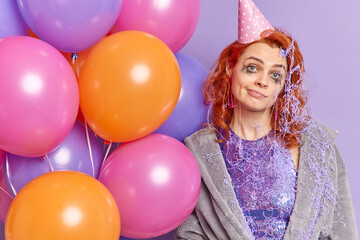 Fototapeta na wymiar Upset redhead young woman smeared with purple cream wears party hat and dressing gown holds multicolored balloons has celebration at home poses indoor. Displeased birthday girl after partying