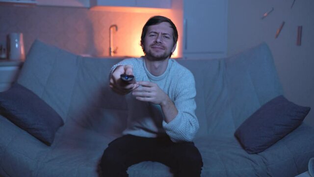 Young bored man sitting on sofa watching television changing the channel with the remote control
