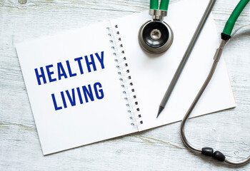 HEALTHY LIVING is written in a notebook on a wooden table next to stethoscope.