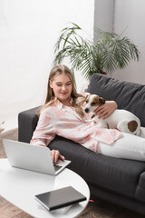 cheerful woman lying on couch with jack russell dog and using laptop in living room