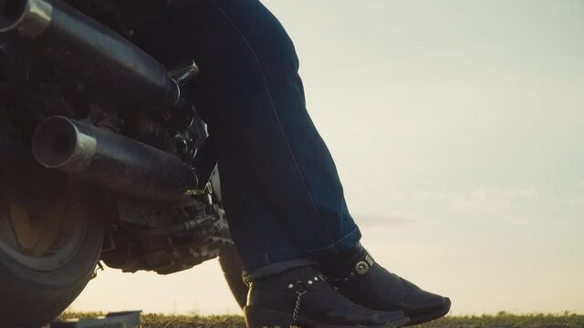 Bottom view of standing motorbike on which motorcyclist leans putting his foot on foot. Closeup of leather biker boots with silver hardware matching motorcycle