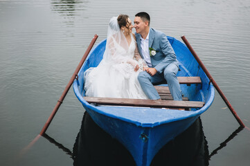 Fototapeta na wymiar Stylish groom in a suit and a cute bride in a white lace dress are sitting in a wooden boat, walking and swimming on the lake, enjoying the beautiful nature. Wedding portrait.