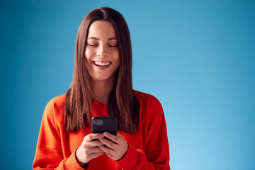 Studio Portrait Of Smiling Young Woman Looking At Mobile Phone Against Blue Background