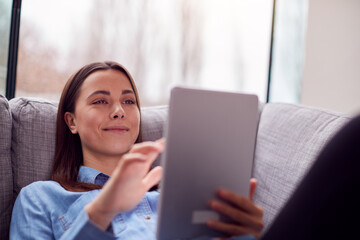 Fototapeta premium Smiling Young Woman At Home Lying On Sofa Looking At Digital Tablet