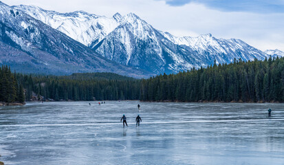 Tourists doing ice-skating in Johnson Lake frozen water surface in winter time. Snow-covered mountain in the background. Banff National Park, Canadian Rockies, Alberta, Canada.