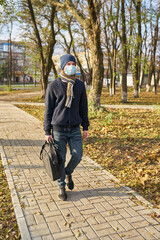 A young guy in a hat with a briefcase is walking down the street to work in a medical mask.