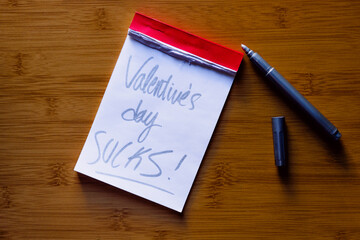 Small notepad with red border on a wooden background with Valentine´s day sucks! written