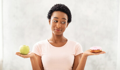 Healthy diet vs junk food. Black lady holding apple and donut in her hands, tempted to eat dessert...