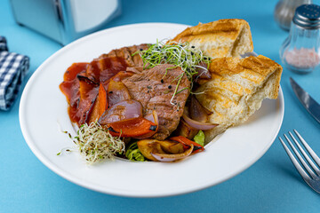 Warm salad with beef, vegetables, croutons and microgreen in a white plate on the restaurant table. Meat food in a bowl. Chop, medallions, cutlets. Blue background, close-up.