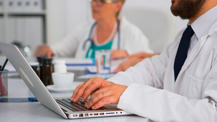 Close up of doctor using laptop, writing treatment informations while coworkers discussing in background during medical conference sitting at desk in hospital office. Team of doctors brainstorming