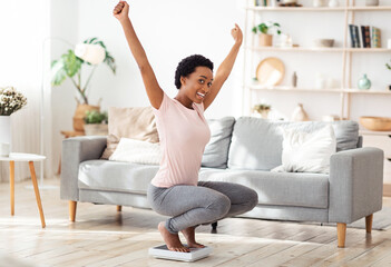 Young black woman sitting on scales, raising hands up in excitement, happy with result of her...