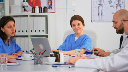 Woman assistent typing on laptop at desk looking at camera and smiling in busy workday in modern medical office. Woman specialist writing on notepad during brainstorming in meeting room