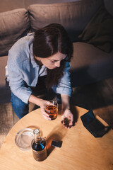 overhead view of woman holding glass of whiskey and coins near bottle and empty wallet