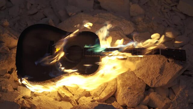 Guitar burning is on fire on stones. Acoustic western guitar burn in darkness. Flame envelopes the guitar