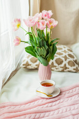 A mug of hot tea, a pink knitted blanket, a bouquet of tulips in a vase, a candle on the bed. Breakfast in bed. Cozy.