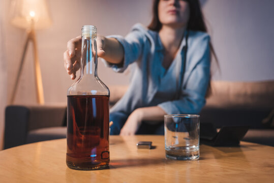 cropped view of alcoholic woman taking bottle of whiskey from table, blurred background