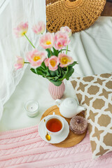 A mug of hot tea, a pink knitted blanket, a bouquet of tulips in a vase, a candle on the bed. Breakfast in bed. Cozy.
